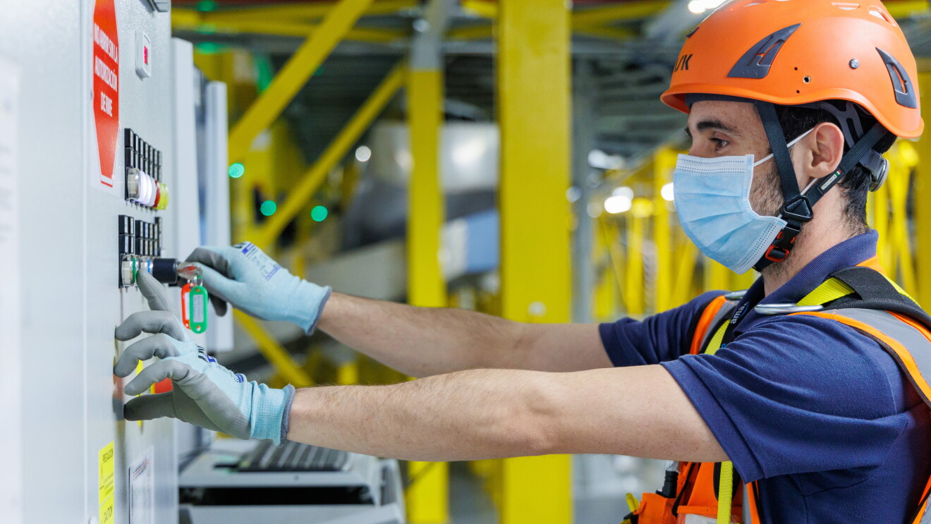En un centro logístico de Amazon aparece un asociado con las manos en unos interruptores. Rodeado de una estructura metálica de color amarillo el empleado va con casco y botas de seguridad, chaleco, camiseta de manga corta azul. 