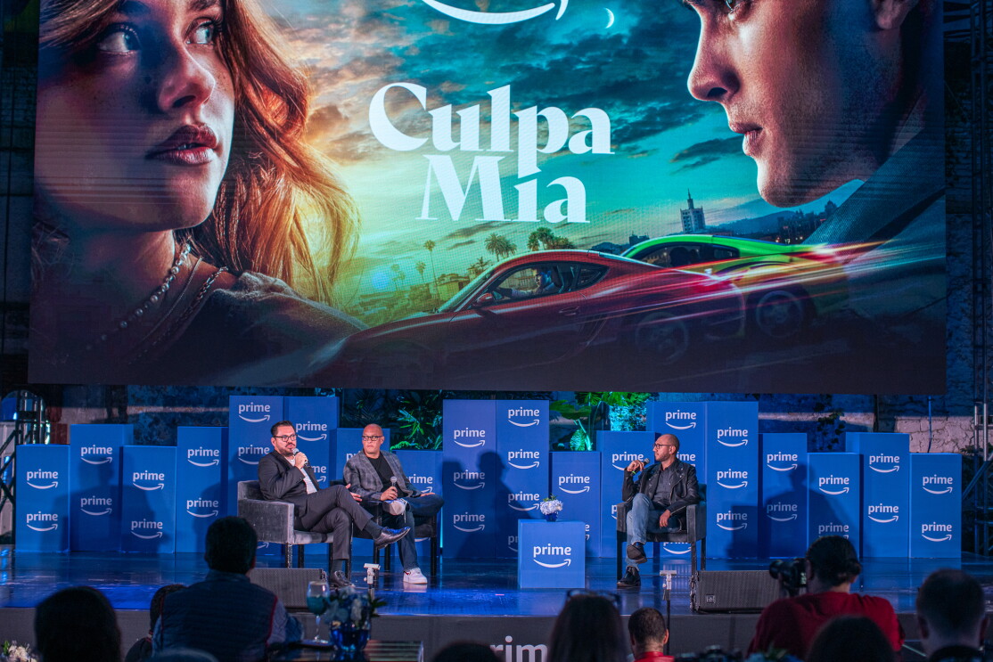 En un escenario con cajas con el logo de Prime, aparecn tres personas sentadas que no se pueden identificar y encima de ellas el cártel de la película Culpa Mía. 