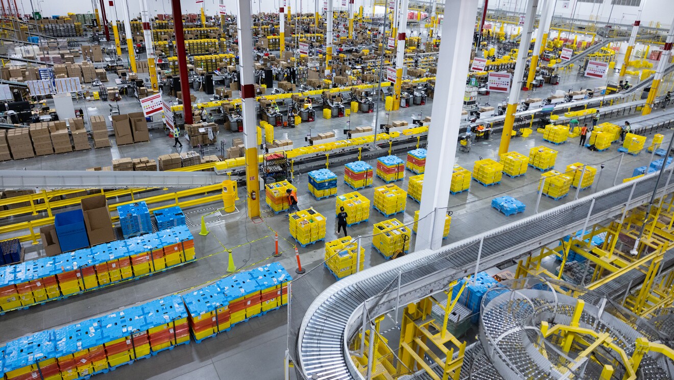 Panorámica de un centro logístico de Amazon. Es una foto sacada desde una altura casi del techo y todo lo que se ve tiene una perspectiva muy amplia. Se ven cintas transportadoras, una parte con cajas amarillas y azules de clasificación y otra parte que son estaciones de preparación de pedidos.