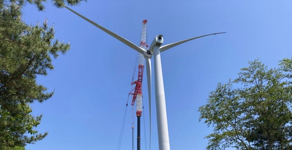青空を背景に、白い風力発電の風車がクレーンで組み立てられている。本体の柱部分に羽を取り付けているところ。左右には緑の木々が見える。
