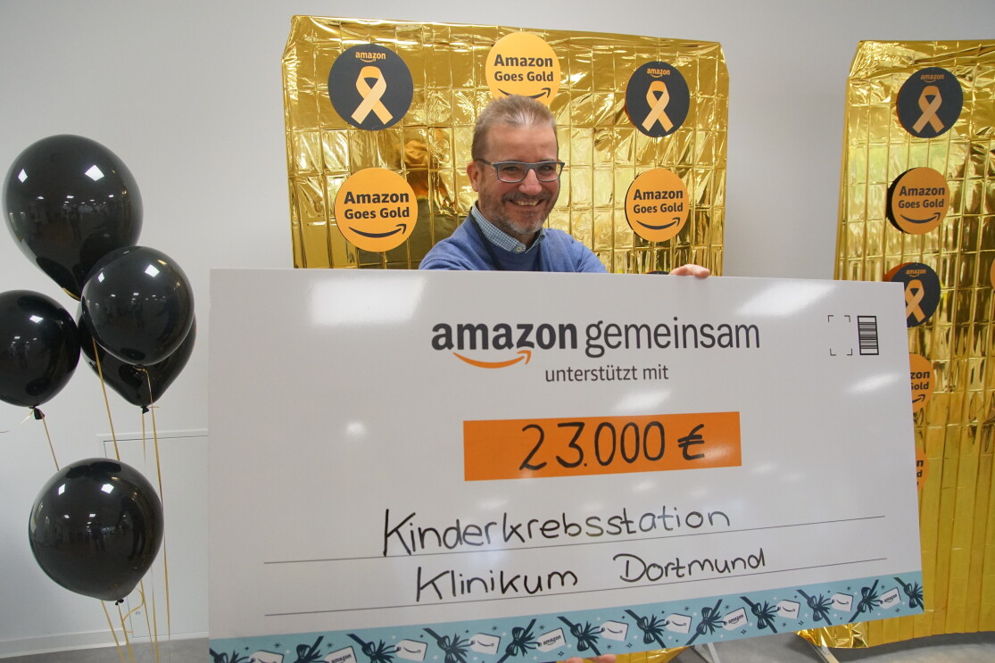Ein Herr mit Spendenscheck vor einem goldenen Fotoleinwand mit Amazon-Goes-Gold-Stickern.