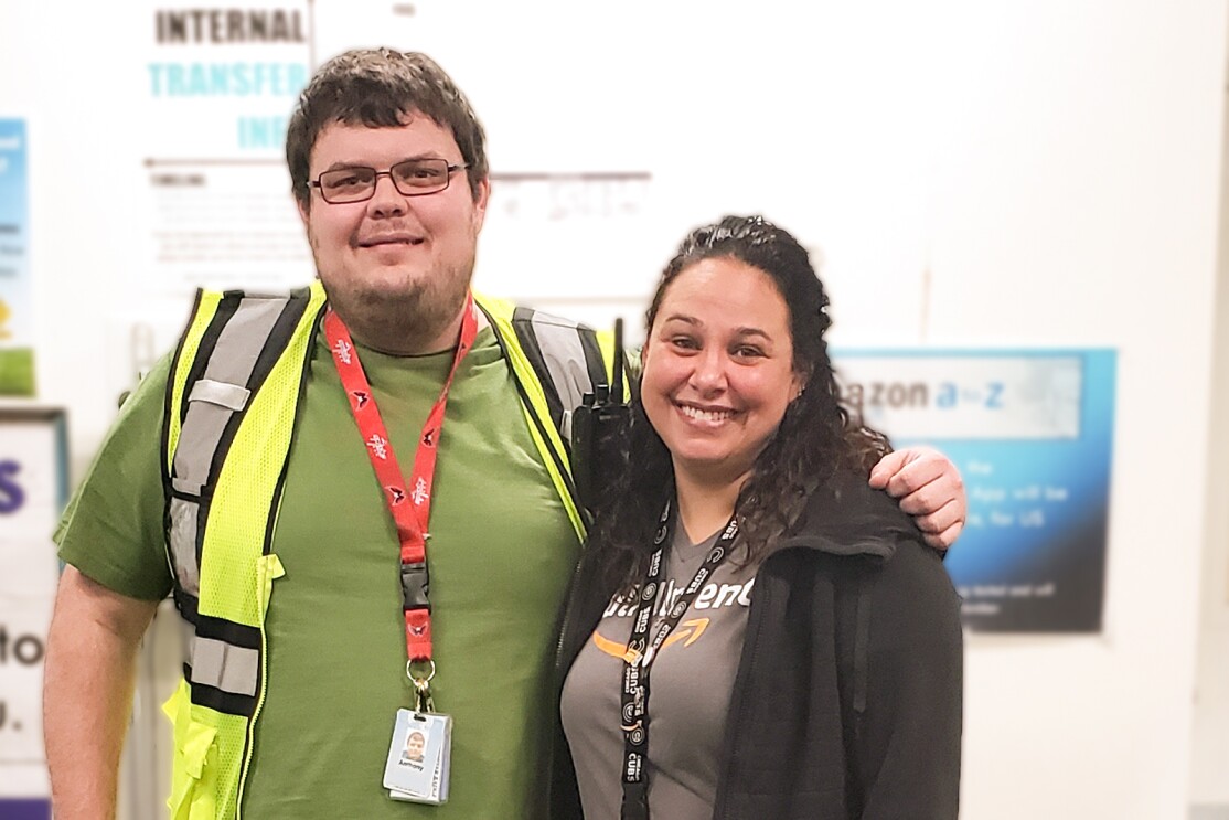 A woman and a man pose together for a photograph. The man wears a green T-shirt and a yellow safety vest. The woman wears a black fleece jacket and a gray T-shirt with the Amazon smile logo.