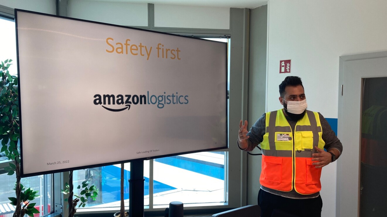 Spezialist für Arbeitssicherheit bei Amazon Mustafa Almujahed bei einer Schulung zu Thema Safety first. Er steht mit einer gelb-orangenen Weste und Maske bekleidet vor einem Schildschirm mit der Aufschrift "Safety first. Amazon logistics".