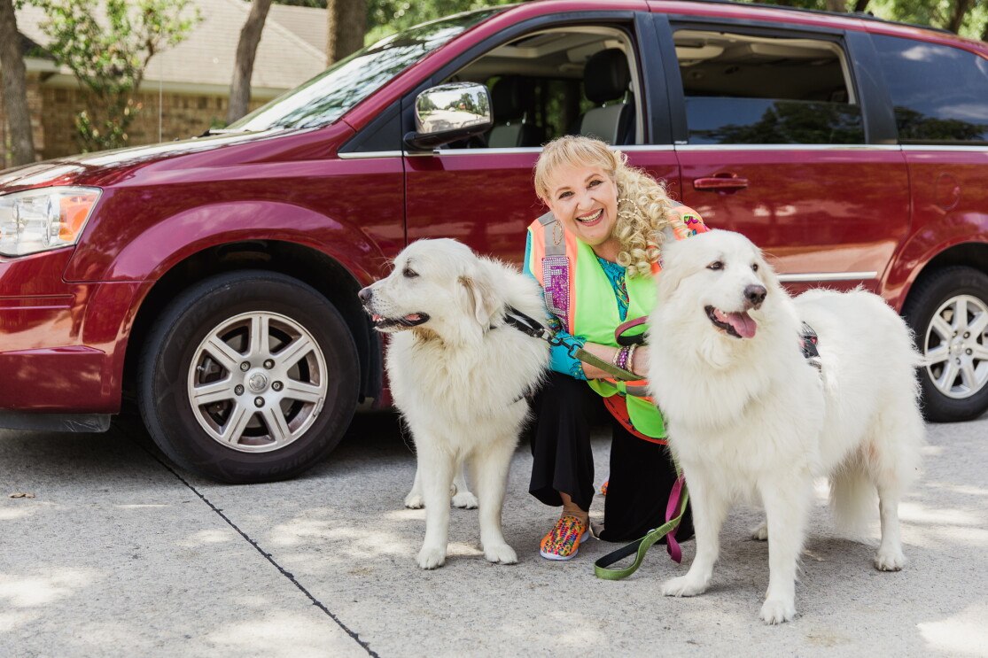 A woman in a bedazzled Amazon Flex vest stands in front of her maroon mini-van. She is smiling at the camera and standing with her two white dogs, who are wearing service animal vests. 