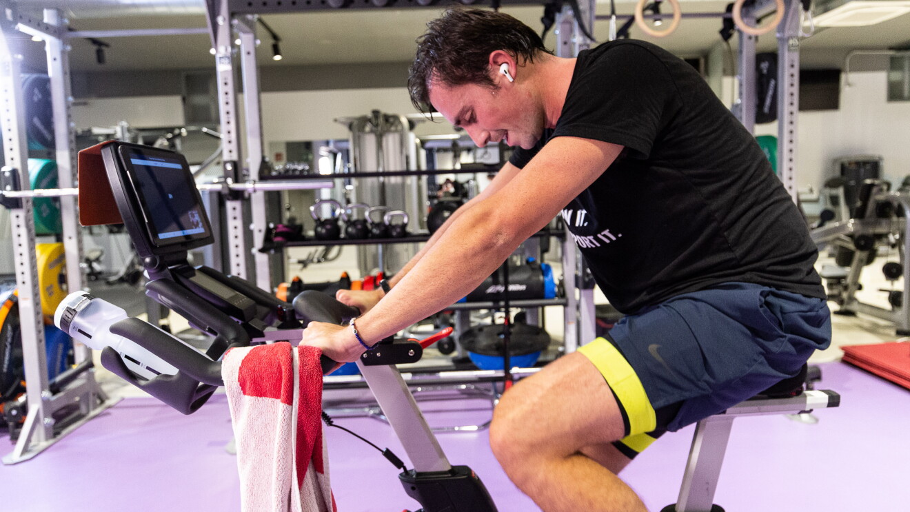 Tiago Moreira-Rato pedaleando para cumplir el reto. En un gimnaso está Tiago montado un bicicleta estática y escuachando música, Tiene un toalla apoyada en el manillar. 