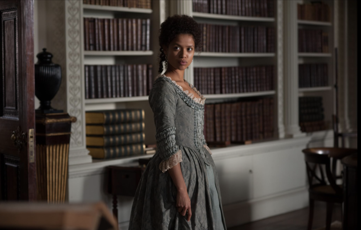 A woman in a dress standing in front of bookshelves, looking at the camera with a smile.