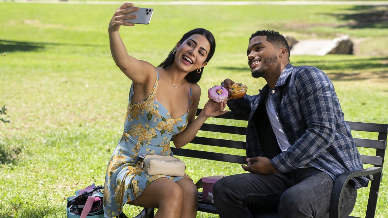 Emeraude Toubia and Rome Flynn are sitting on a bench in a park taking a selfie while holding doughnuts.