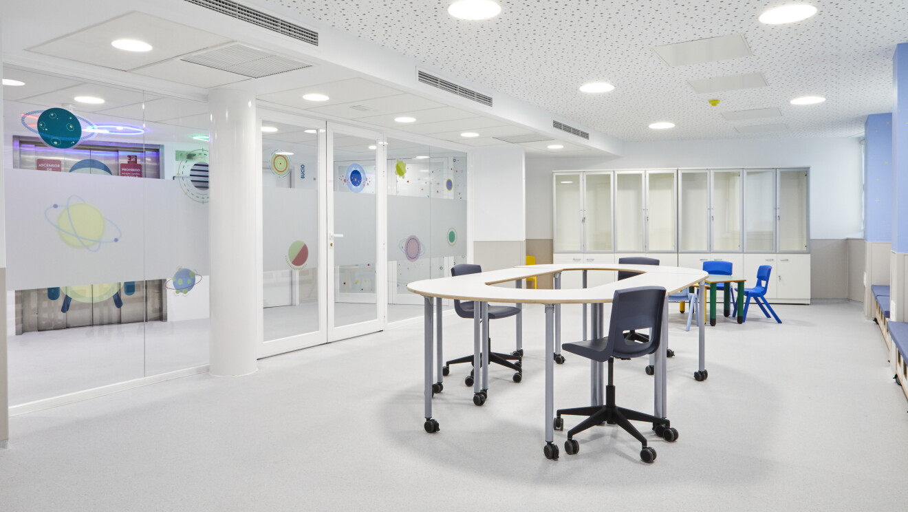 Aula formación de futuros astronautas del proyecto La Paz en Órbita del Hospital Universitario de La Paz. Una sala rectangular con una mesa redonda blanca con tres sillas grises con ruedas. Al lado hay otra mesa verde para pequeños con tres sillas de plástico de color azul. Al lado derecho una pared de color morado. De fondo cuatro estanterías blancas con la parte superior con cristales y la inferior con puertas. La pared de la izquierda es de cristal con vinilos que tienen dibujos de planetas y estrellas. El techo es blanco con estrellas de color gris.  