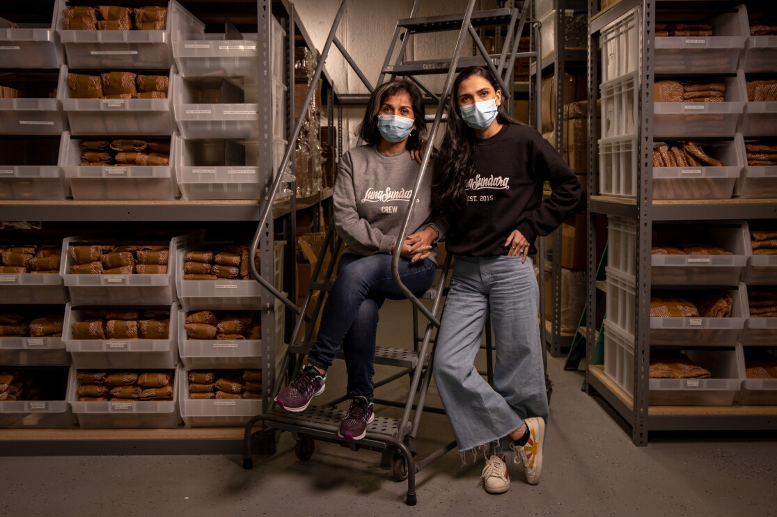 Two women in sweatshirts and jeans flanked by shelving in a warehouse space.