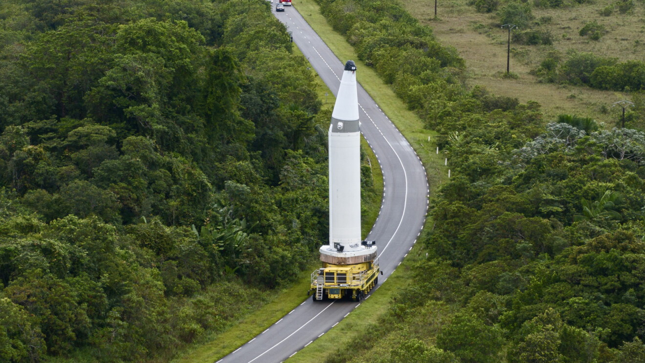 Large industrial truck transporting a large white spacecraft on a winding rural highway