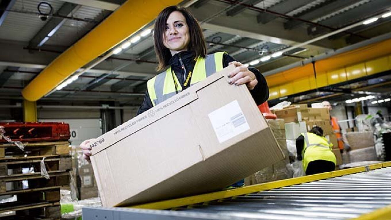 Amazon Mitarbeiterin stehe am Fließband und hat einen Karton in der Hand.
