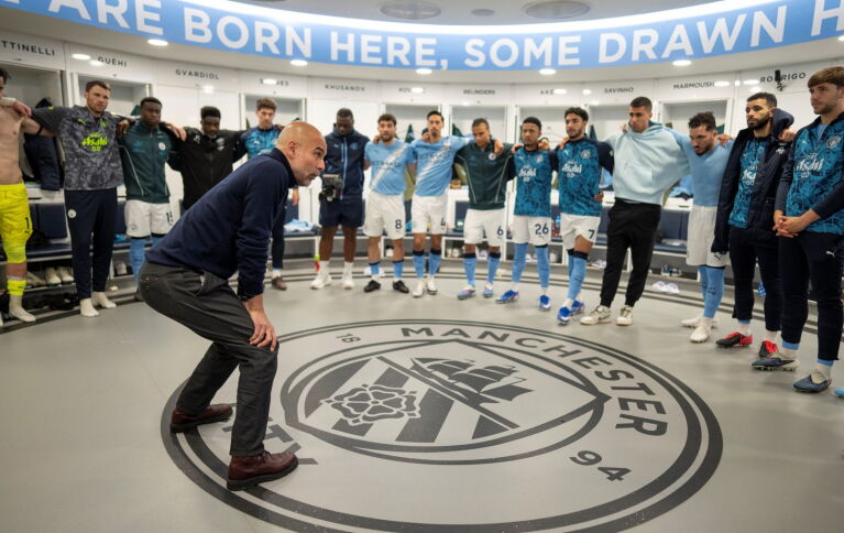 Pep Guardiola stands in the Man City dressing room, giving his players a team talk 