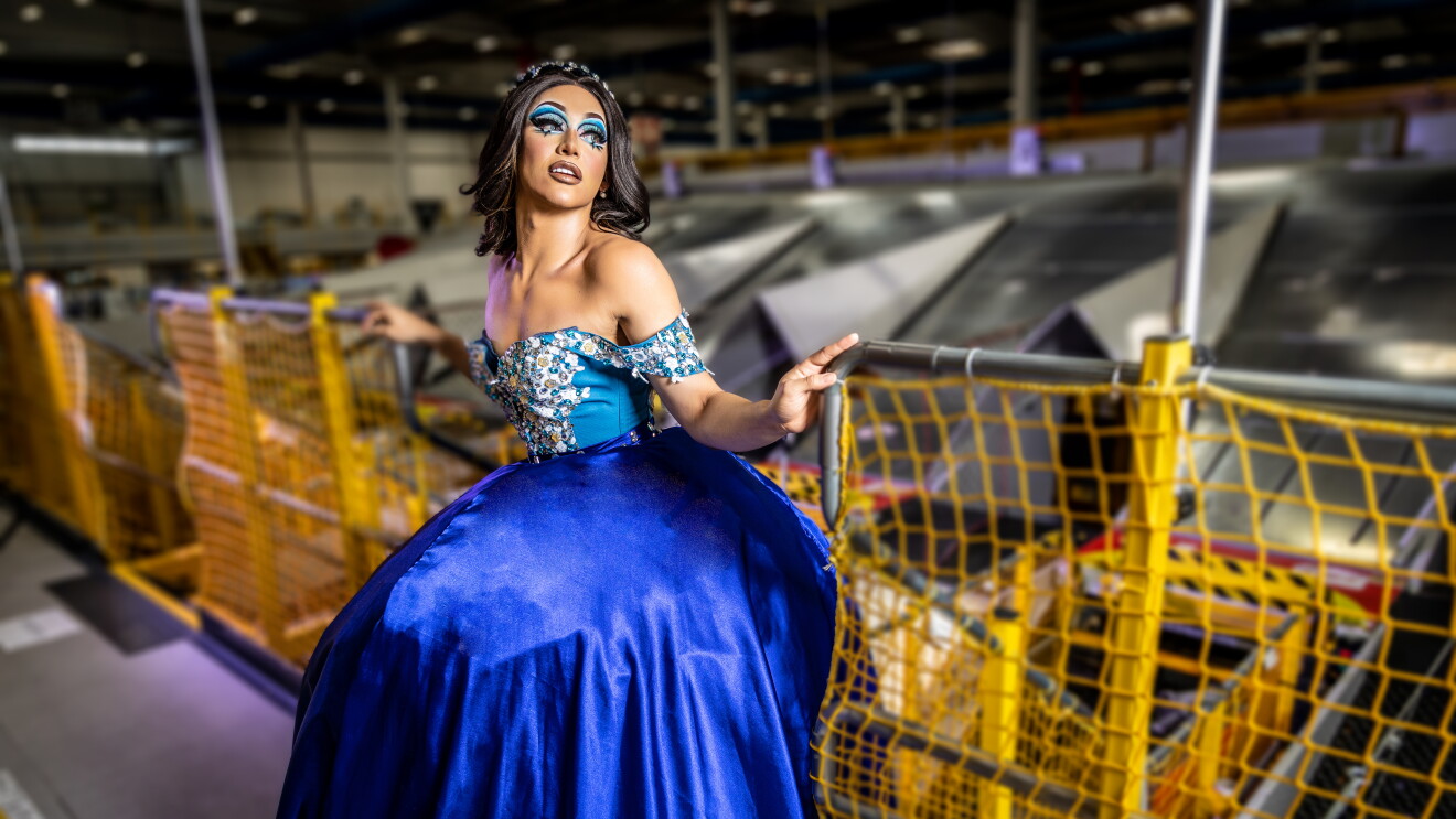 Rey_Mariana Stars. Es una drag queen que trabaja en Amazon. Va con un vestido de fiesta. La parte superior es un corsé azul con pedrería y la parte inferior es azul más oscuro y con muchísimo volumen. Tiene el pelo marrón oscuro y largo hasta la cintura. Lleva una diadema. Los ojos maquillados también de tonos azyules. Mariana está apoyada a una barandilla de seguridad de Amazon de color amarillo y de findo se ve el tejado de una parte de un centro logístico. 
