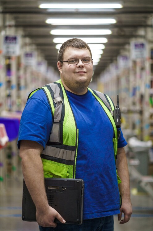 A man poses holding a laptop computer. He wears glasses, a blue T-shirt, and a yellow safety vest.