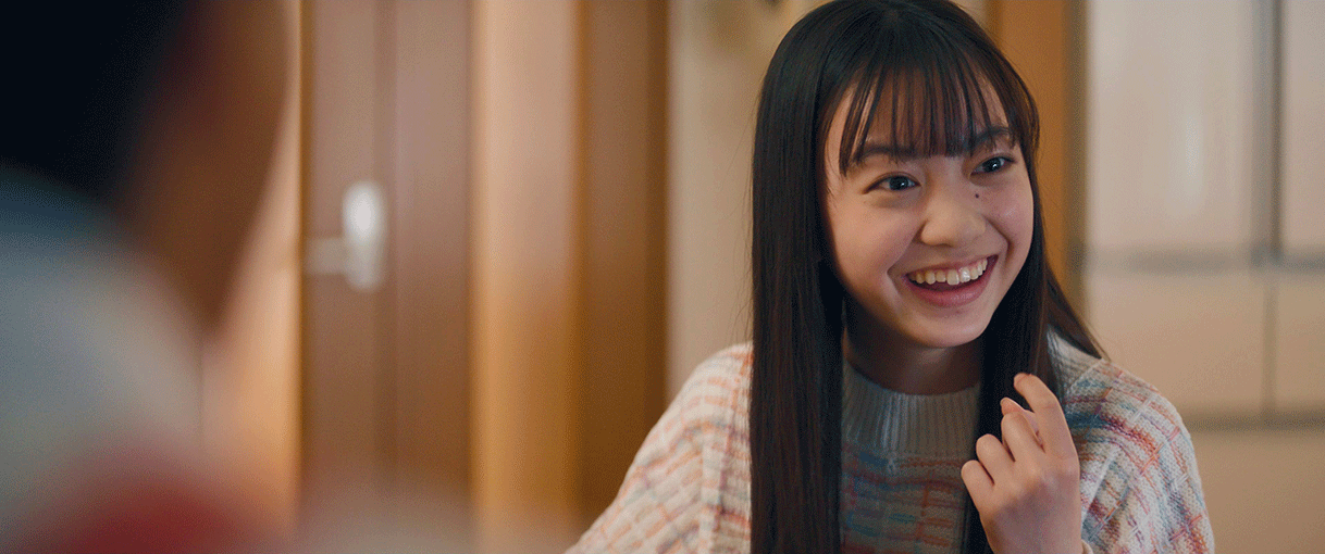 落ち着いた色調の室内、セーターを着た黒髪ロングの少女が右の方を見て笑っている。左側手前にもう1人人物がいるが、ピントがぼやけている。