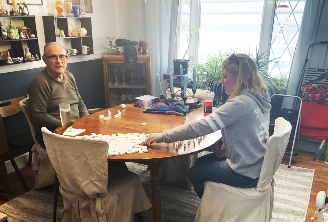 An image of a man and a woman playing dominoes at a kitchen table. The man is looking at the camera and smiling.