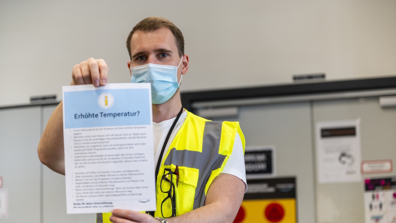 Ein Mann in gelber-Sicherheitsweste und Mundschutz hält ein Informationsblatt mit dem Titel "Erhöhte Temperatur" hoch