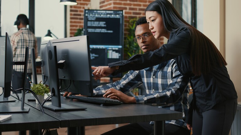 A man is sitting at his desk, his coworker is standing next to him, reviewing content on his desktop computer.