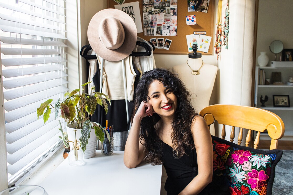 An image of a woman smiling for a photo while resting her head on her hand. She is at a desk and there is a bulletin board with various items including a wide-brim hat and photos behind her.