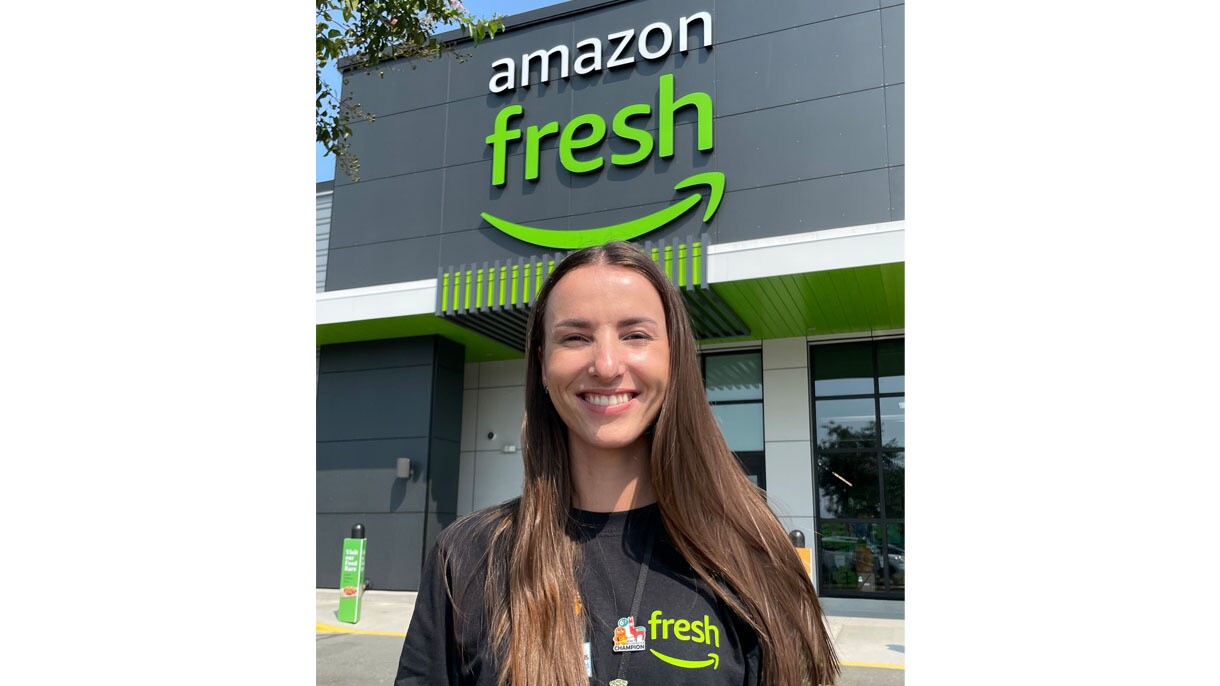 A headshot image of a woman standing outside of an Amazon Fresh store smiling for a photo while wearing her Amazon Fresh shirt and a black lanyard with an Amazon pin on it.  