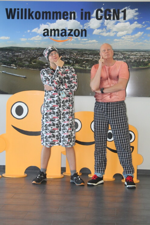 Zwei Mitarbeiter sind vor einer Fotorückwand zu sehen: Einer der beiden trägt ein Nachthemd mit passender Schlafmütze, der andere eine karierte Pyjama-Hose mit einem Shirt, auf dem eine männliche Bodybuilder-Brust zu sehen ist