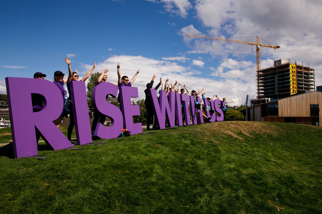 "Rise with us" sign for the Special Olympics USA Games, set on a hillside in Seattle. The purple letters are shown with more than a dozen Amazon employee volunteers waving and smiling, as they welcome athletes to the closing ceremony.
