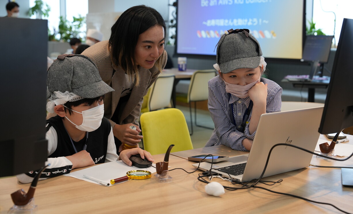 シャーロックホームズのような帽子をかぶり、画面に向かう子どもたちとボランティア。机にはパイプやルーペもおかれている