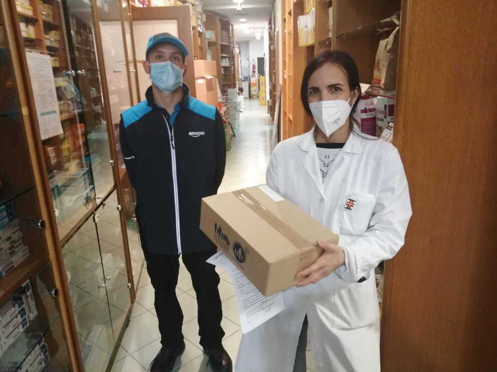 Un autista Amazon consegna un pacco di medicinali ad una farmacista, che indossa un camice bianco. 
