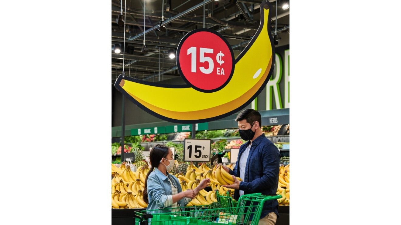 A father and his daughter shop for bananas at Amazon Fresh. Above them is a sign in the shape of a banana with 15 cents above it.