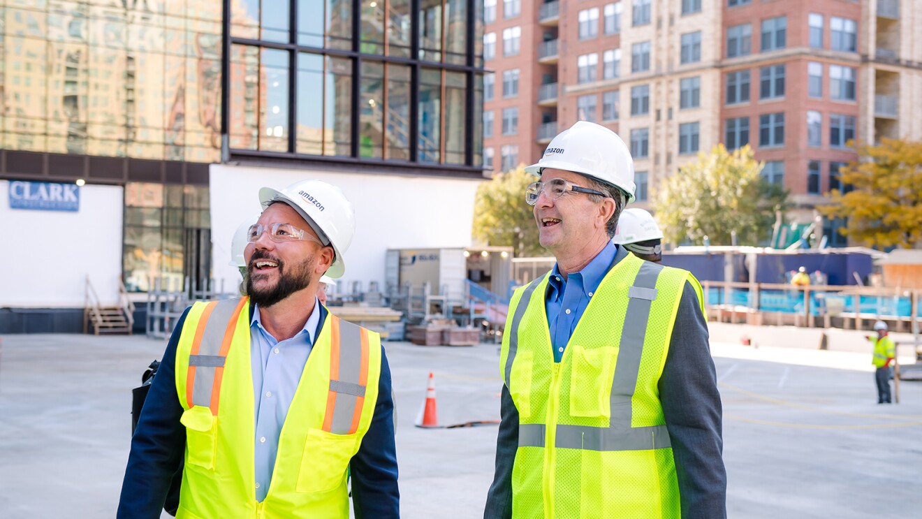 An image of people touring the construction site of Amazon's second headquarters while wearing hard hats that say "Amazon" and bright yellow safety vests.