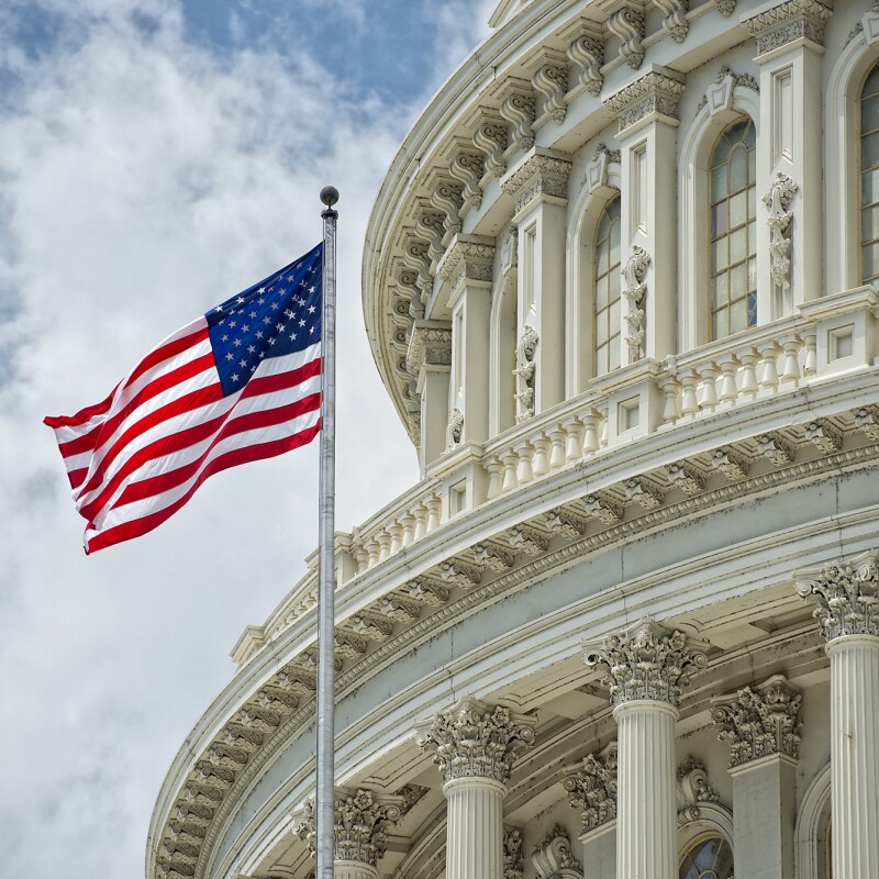 The United States Capitol, against a bright blue sky with fluffy clouds. A U.S. flag flaps in the wind in the foreground. 