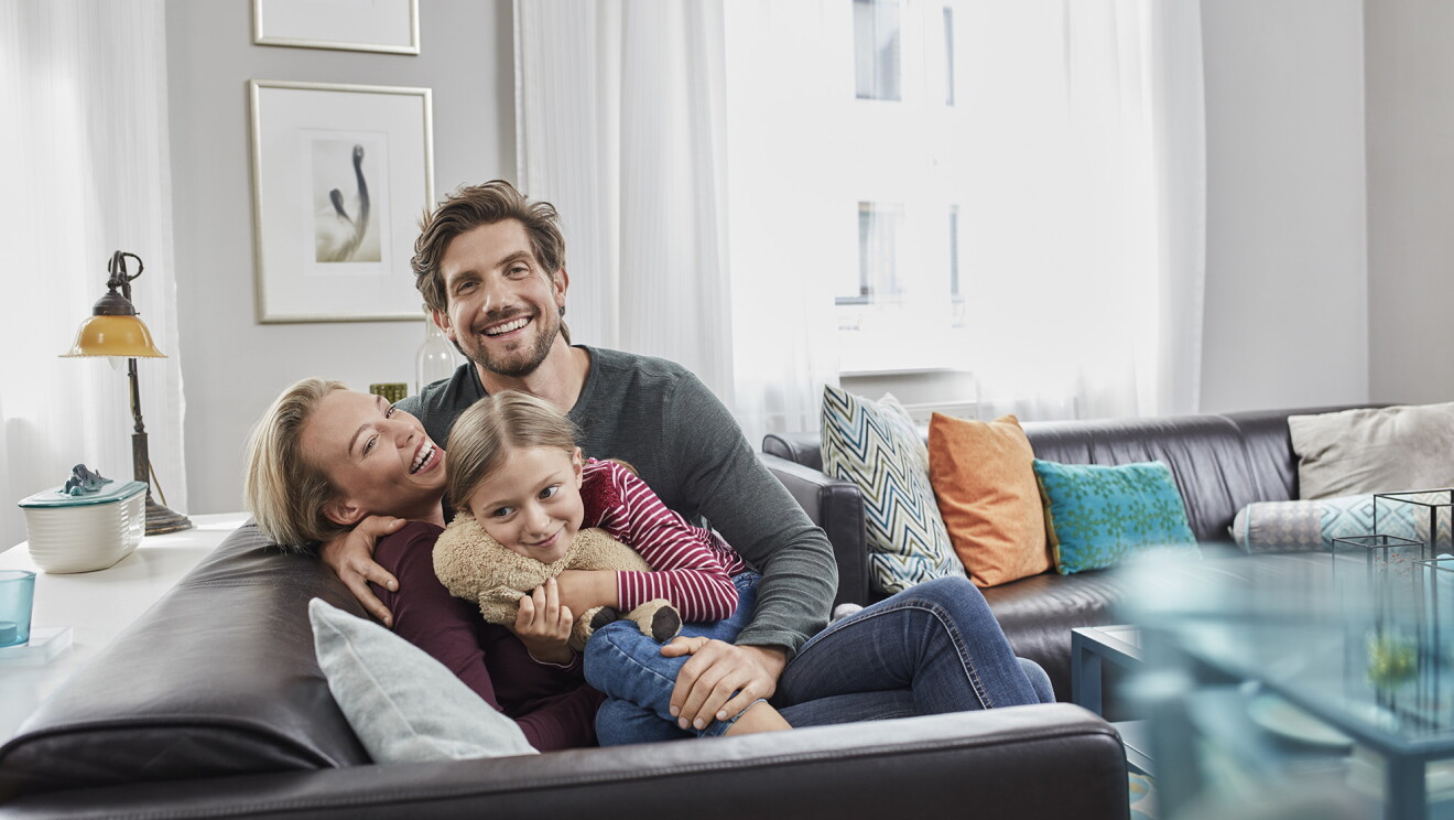 A family of three (mom, dad, child) is seated on a sofa in a contemporary living room. 