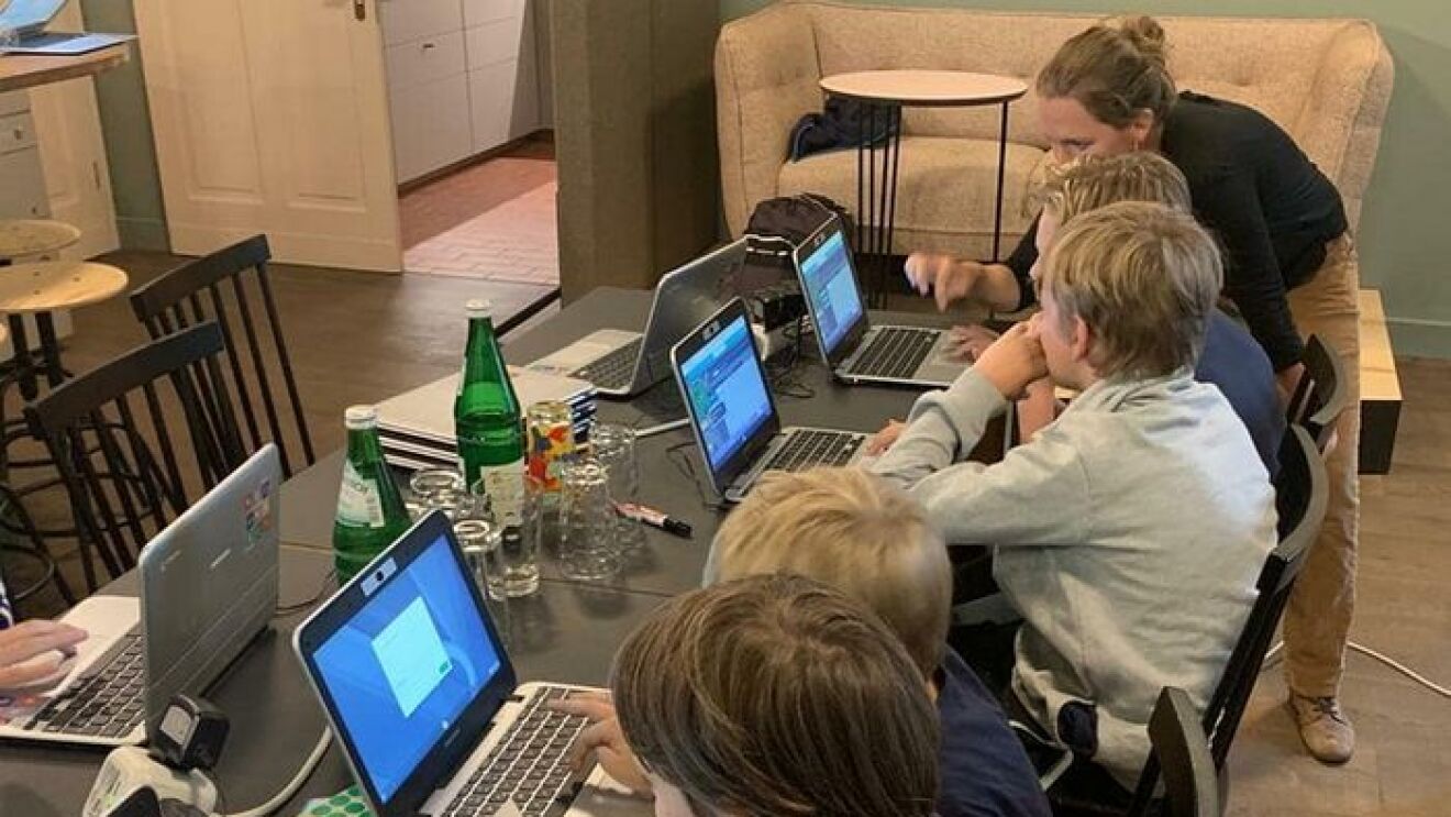 digital.engagiert_oderdojo_Kinder sitzen an einem langen Tisch, vor ihnen stehen Laptops. Eine Frau erklärt ihnen etwas.