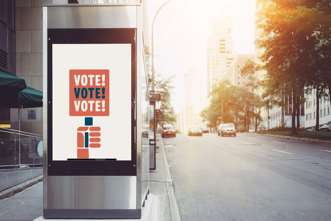 A metal bus station on a city street, with a illustrated poster on it tyat says "vote! vote! vote!" 