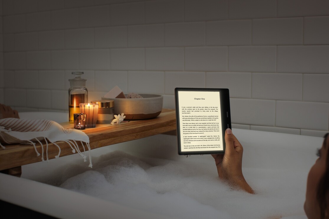 A woman relaxes in a bubble bath while reading a Kindle Oasis in candle lighting.