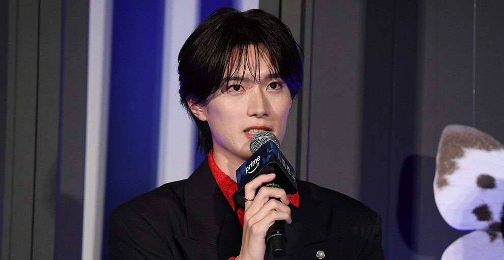 黒と白、青の壁を背に微笑む青年のクローズアップ。黒い上着に赤いシャツを着用。