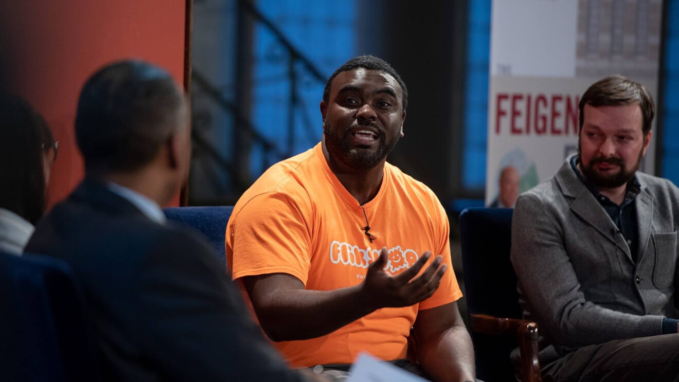 一个男人对一群人讲话的画面。他穿着一件橙色衬衫,上面写着“Flikshop”。