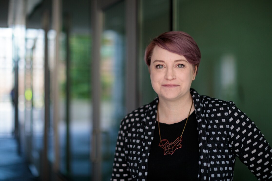 A woman in a black and great patterned jacket looks at the camera.  She's wearing a necklace with a geometric design. 