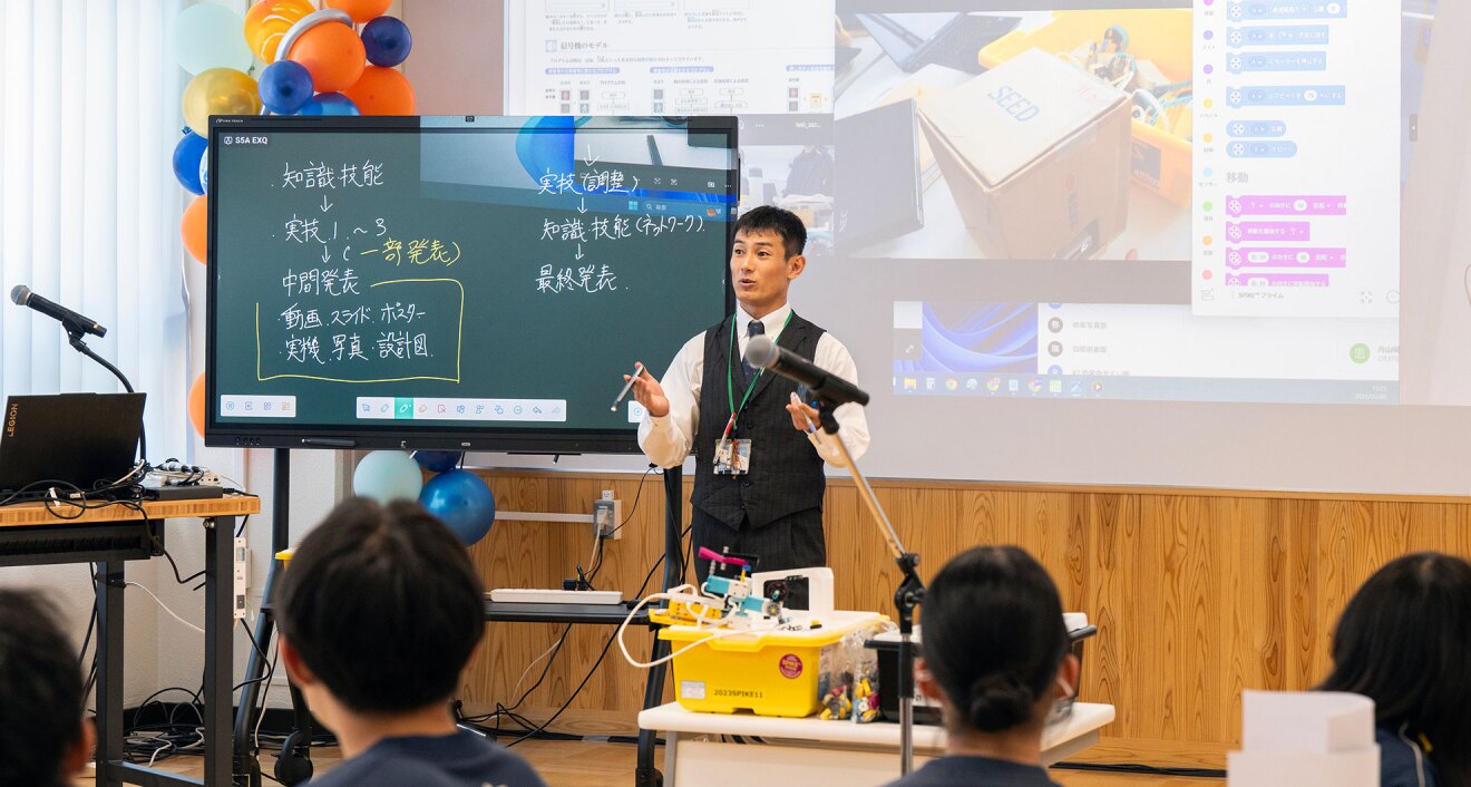 教室で、大型のデジタルディスプレイとプロジェクター投影画面を背にして立つ教師。手前には教師の話を聞く生徒たちの頭部が見える。ディスプレイの後ろに風船の装飾。電子ホワイトボードには「実技」や「発表」などの文字で工程が表示されている。投影には箱の画像やチャット画面が見える。