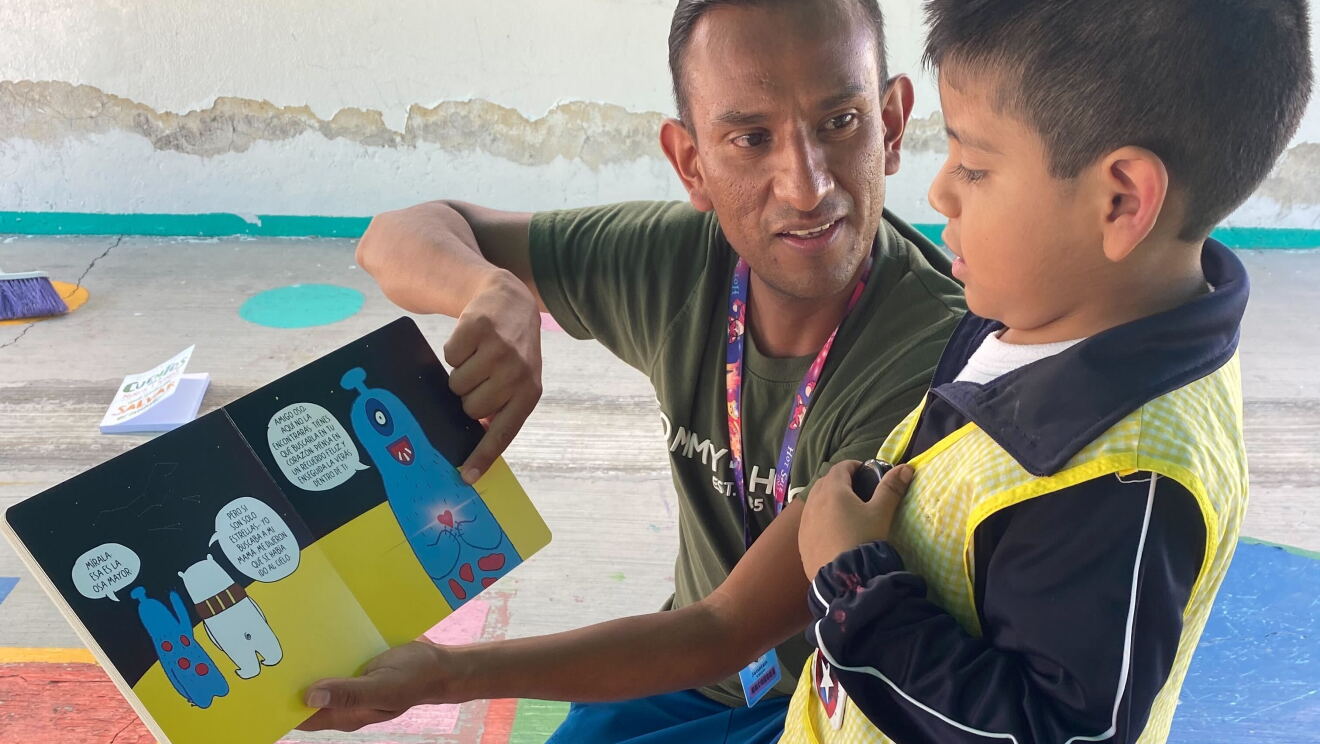 En un patio de colegio, un voluntario de Amazon con dos niños explicándoles un cuento. 