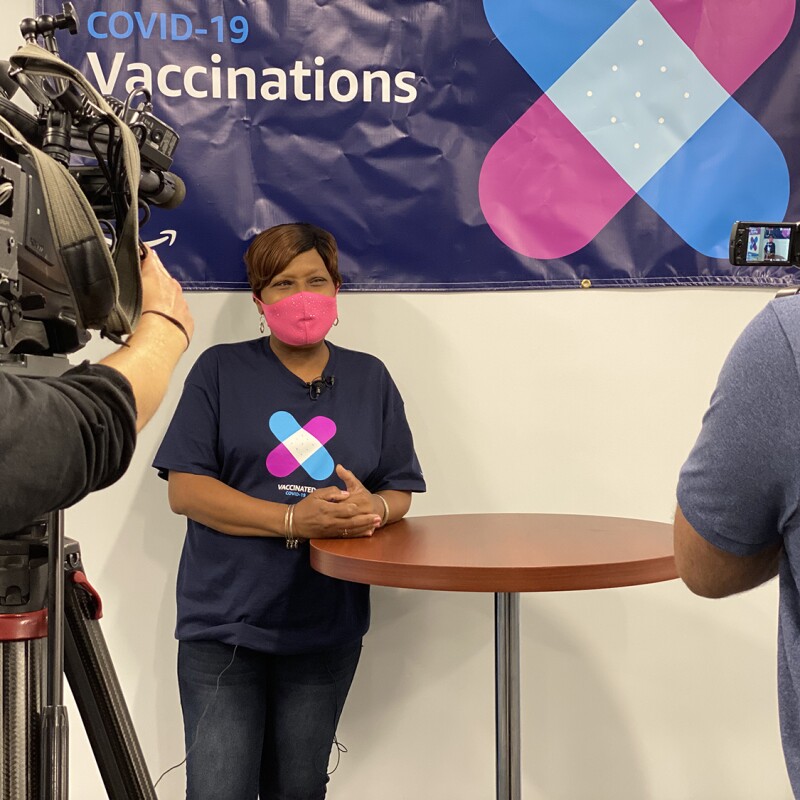 An image of a woman wearing a pink mask while smiling and leaning against a tall table. She is wearing a shirt showing she has been vaccinated and being filmed by a film crew. 