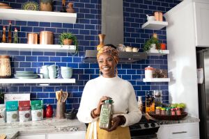 sylvia kapsandoy the founder of usimplyseason stands in front of a kitchen stove and countertop holding a carton of spice seasonings