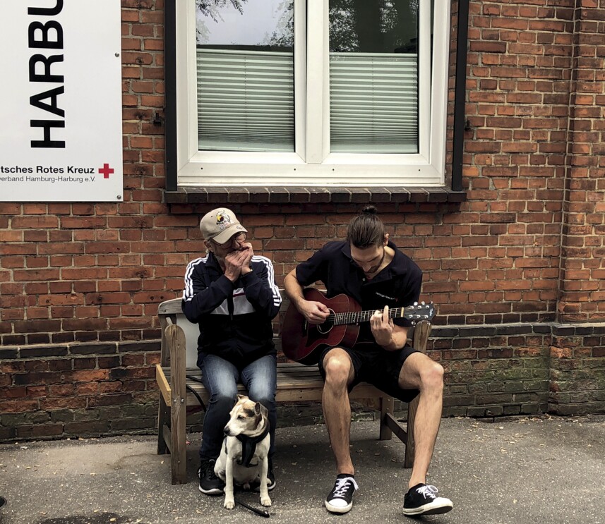 Ein jungen Mann sitzt auf der Bank vor einem Haus und spielt Gitarre. neben ihm sitzt ein älterer Mann und spielt Mundharmonika.