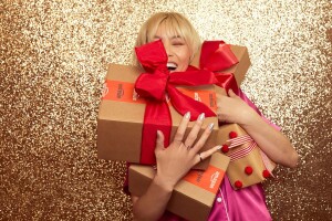 An image of a woman with blonde hair holding Amazon boxes in front of a gold backdrop