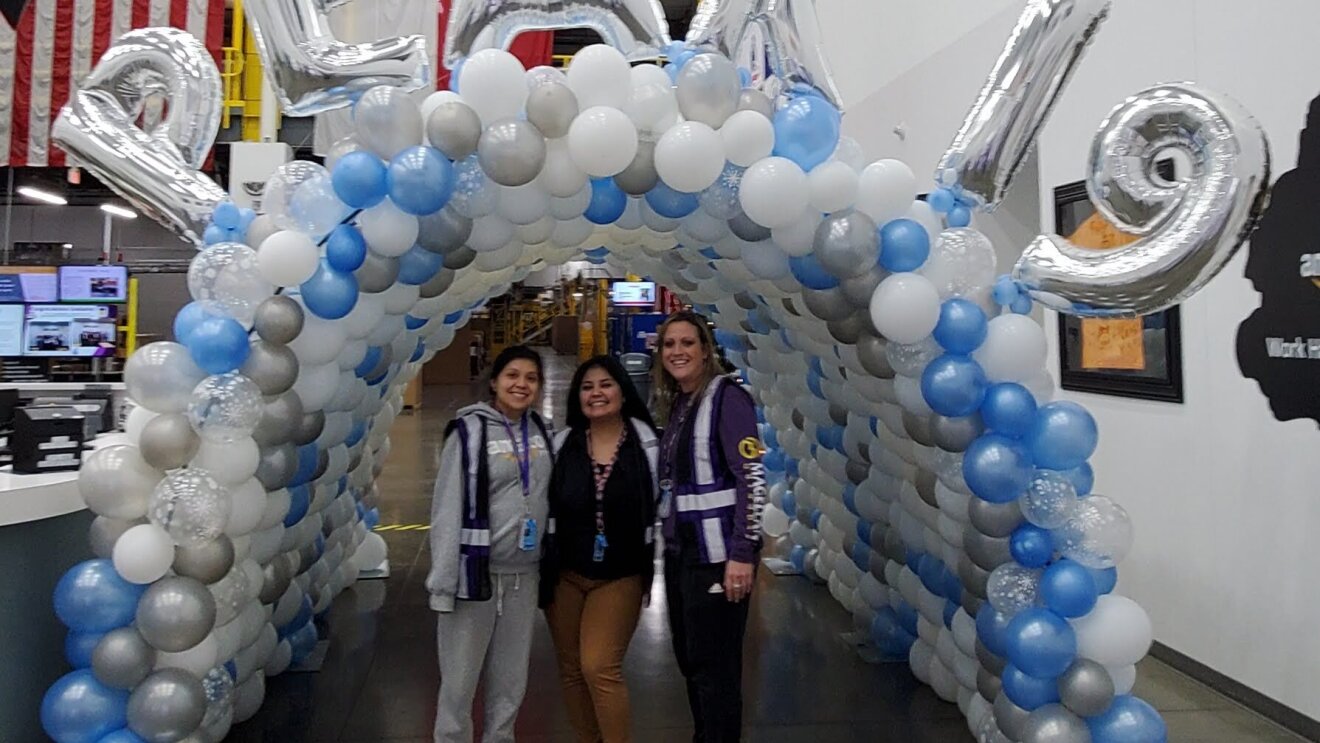 Three Amazon associates stand under a balloon bridge with "PEAK 19" balloons displayed on the top.