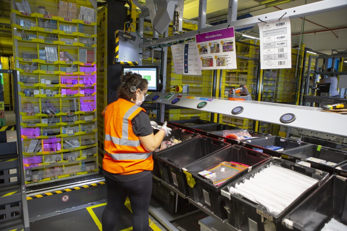 En un centro logístico de Amazon, de izquierda a derecha, aparece: una estantería amarilla llena de productos. Una trabajadora de Amazon de espaldas a la cámara. Lleva pantalón y camiseta negra, chaleco de color naranja y pelo rizado por el hombro. Está agarrando un producto con las dos manos. Se encuentra delante de una estación con cajas negras llenas de productos.