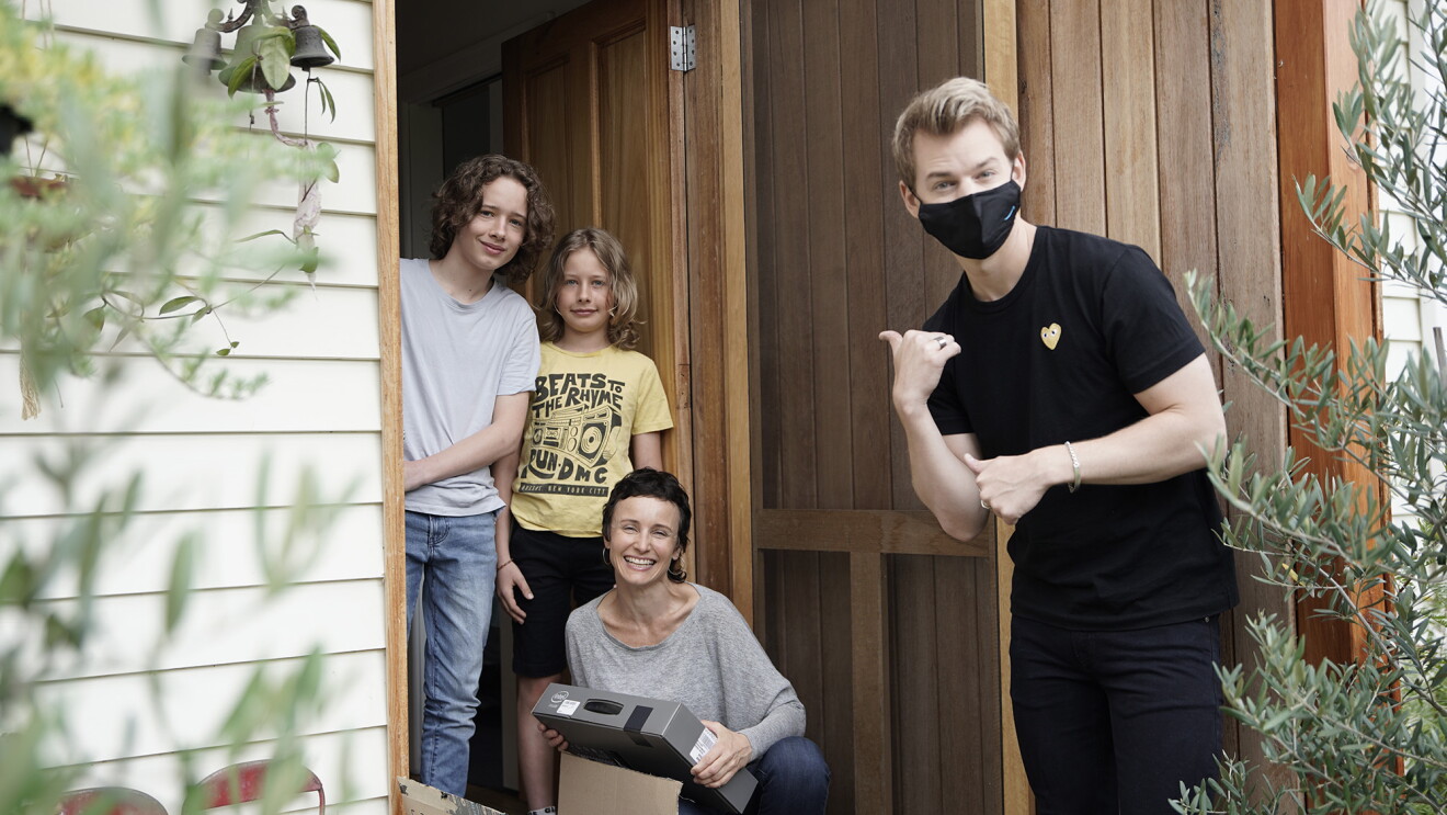 A man stands by the front door of a home, he wears a black mask and black outfit, looking at the camera, he has his hand in a gesture that suggests "look at that / them," toward a family smiling at the camera. 
