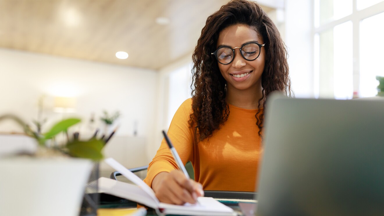 An image of a woman wearing an orange shirt and glasses. She is writing on a notepad with a laptop in front of her. 