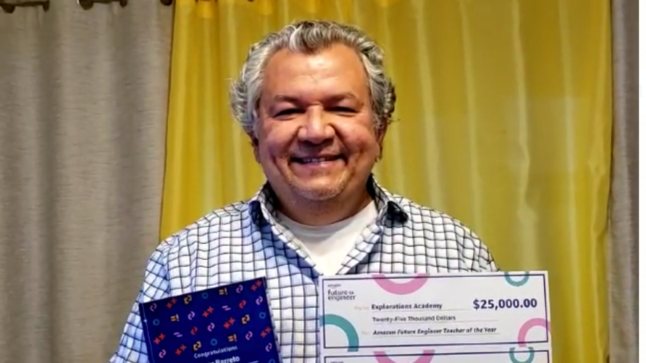 A man smiles for a photo holding a paper that says he won the Amazon Future Engineer Teacher of the Year award and two checks--one for $25,000 and the other for $5,000.