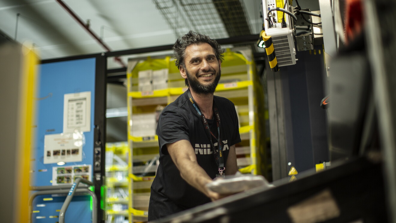 Elia works in a FC. He has a black tshirt, a big smile and he is picking an item. ELIA VILLARDITA - Operatore di magazzino :"Sicurezza sul lavoro significa creare un ambiente lavorativo dove non ci sono condizioni di pericolo. Avere sicurezza sul lavoro porta tranquillità, è bello sapere che i rischi sono quasi nulli, questo ci permette di lavorare meglio e quindi in modo più produttivo".